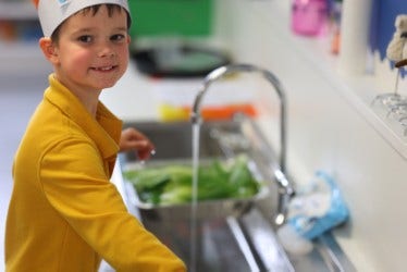 Boy student at sink washing vegetable produce ready to use for the Kindy Cafe.