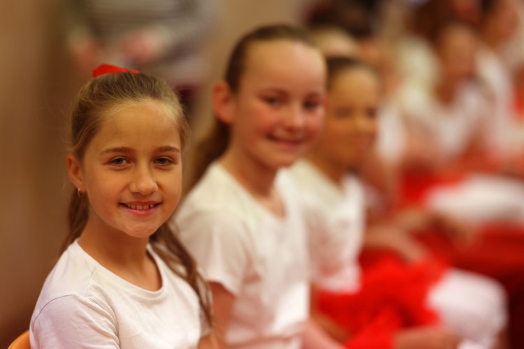 Girl students dressed in white T shirts smiling.They are getting ready to perform with the dance ensemble at our school's open day assembly in the school hall.