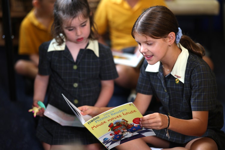 A year 6 female student reading a book to a female kindergarten student. They are sitting on the floor. The kindergarten is very engaged in the story and the year 6 student is reading with a smile on her face.