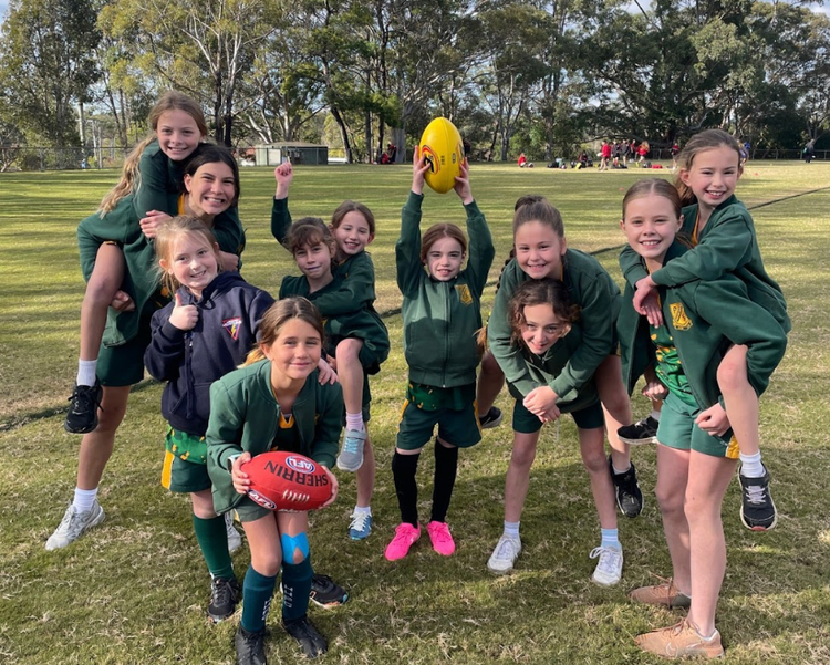 A group of 11 girls who are part of a girls AFL team. All are smiling, with some girls being piggy backed and others with their thumbs up or with hands in the air.